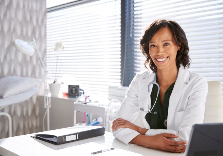 Portrait Of Smiling Female Doctor Wearing White Coat With Stethoscope Sitting Behind Desk In Office