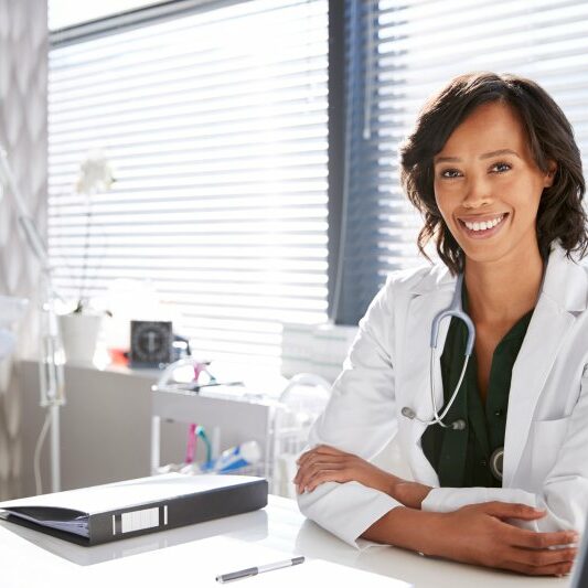 Portrait Of Smiling Female Doctor Wearing White Coat With Stethoscope Sitting Behind Desk In Office