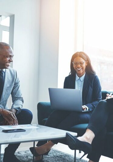 Diverse group of smiling businesspeople talking together while sitting on a sofa in a bright modern office